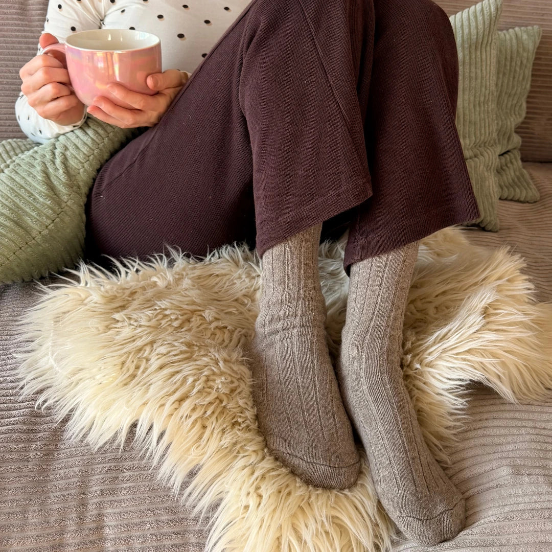 Frau (40+) sitzt gemütlich auf der Couch mit einer Tasse Tee und trägt hellbraune Premium Alpakasocken